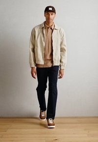 Lys beige bomberjakke, brun ribbestrikket skjorte under, marineblå bukser og brune sneakers med hvite såler. Enkelt design og avslappet passform.