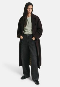 Lange donkerbruine jas over een lichtgroene t-shirt, gecombineerd met brede zwarte jeans. De outfit heeft een comfortabele pasvorm en soepele texturen.