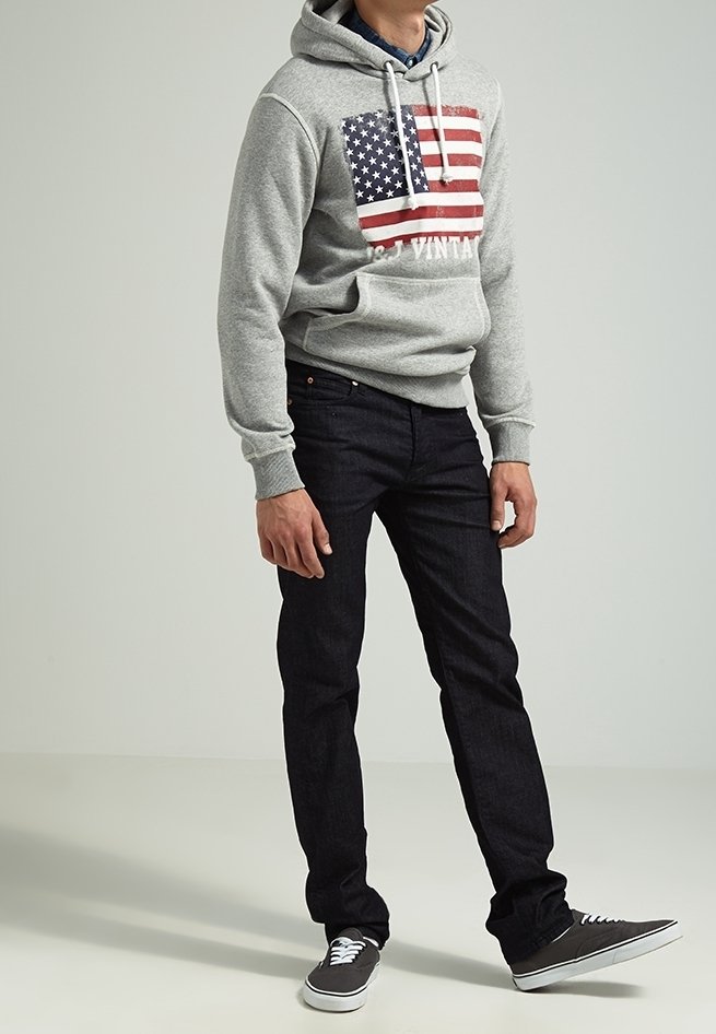 Felpa grigia con grafica della bandiera americana e testo, abbinata a jeans denim scuri e sneakers nere con lacci bianchi. Outfit casual.