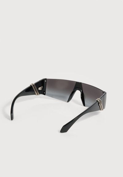Lunettes de soleil enveloppantes noires avec une seule lentille teintée foncée et des accents d'articulation argentés sur les branches, reposant sur une surface blanche.