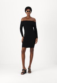 Abito a coste nero con scollo off-the-shoulder, maniche lunghe, vestibilità aderente. Indossato con sandali neri con tacco grosso e dettagli metallici.