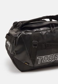 Sac de sport noir en tissu imperméable, avec des sangles réglables, un zip sur le dessus et un logo "TERR" bien visible sur le côté.