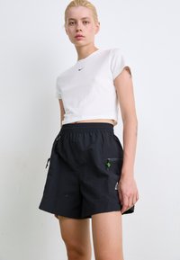 Black shorts with an elastic waistband, side pockets, and a small green accent. The model wears a white cropped T-shirt with a black logo.