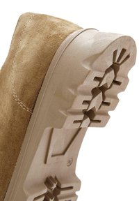 Chaussure en daim beige avec une semelle en caoutchouc texturée présentant un motif strié. Couture visible le long des bords supérieurs de la chaussure.