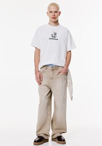 Ung person med kort, blondt hår iført en hvid T-shirt med Honda-logo, beige vidde bukser, mørke sko samt en halskæde og armbånd.