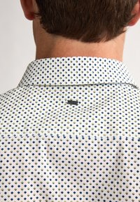 Camicia bianca con un motivo a losanghe blu e verde, dotata di colletto button-down e di un piccolo logo metallico vicino al collo.