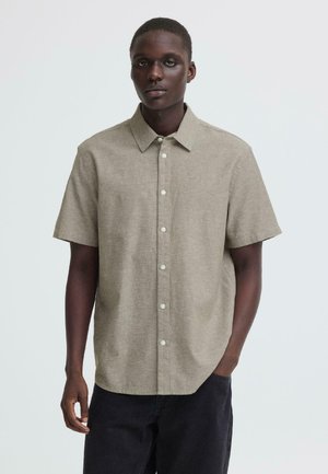 Junger Mann, der ein kurzärmeliges beiges Hemd mit Knöpfen und schwarze Hose trägt, mit einer Hand in der Tasche vor einem schlichten Hintergrund stehend.