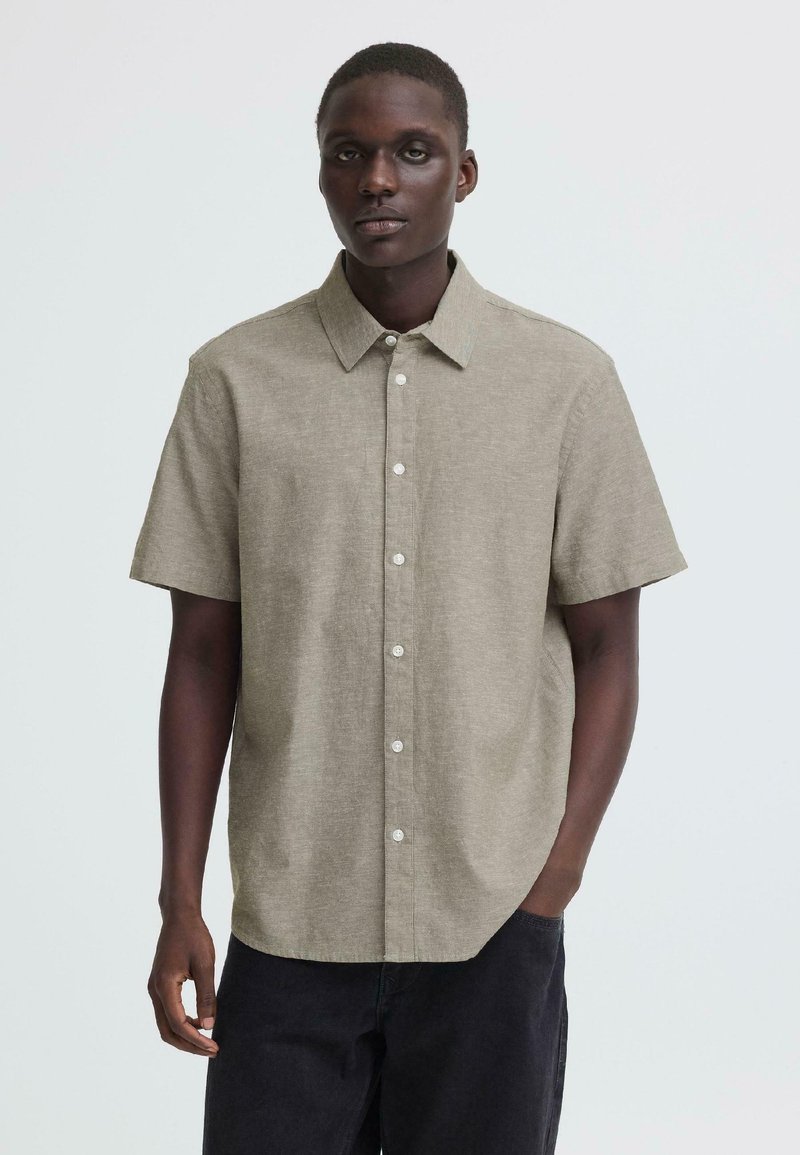 Νέος άνδρας που φοράει κοντομάνικο μπεζ πουκάμισο με κουμπιά και μαύρο παντελόνι, στέκεται με το ένα χέρι στην τσέπη μπροστά σε απλό φόντο.