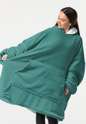 Frau in übergroßer tealfarbener Kapuzen-Fleece-Decke mit großer Fronttasche, die die Seiten hält, während sie lächelt und nach rechts schaut.