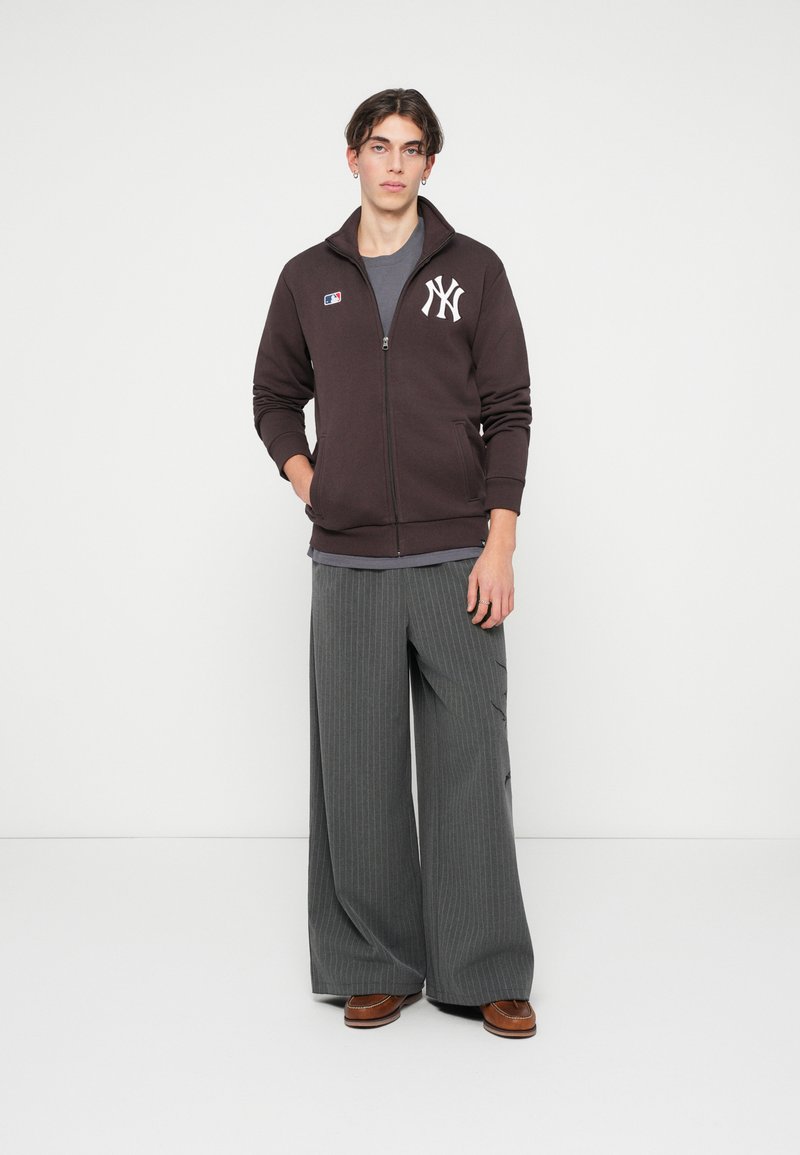 Brun zip-up hoodie med hvidt logo, grå t-shirt under, grå stribede vide bukser og brune sko. Minimalistisk, afslappet design.