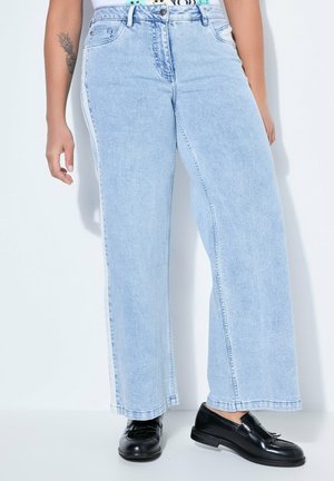 Person trägt hellblaue Jeans mit weitem Bein und weißen Seitenstreifen sowie schwarze Lederloafer, steht vor einem schlichten Hintergrund.