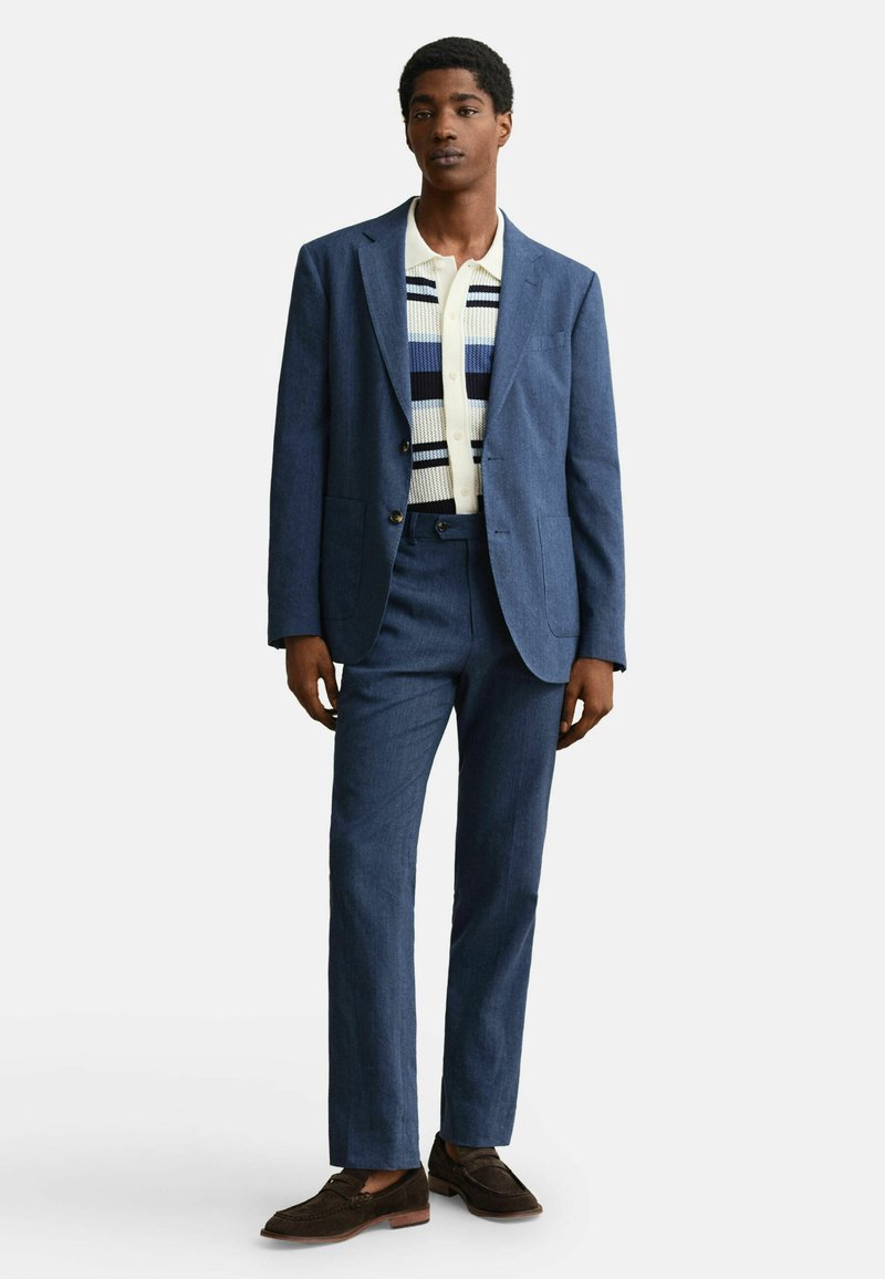 Hombre con traje azul, suéter de punto a rayas y camisa crema, usando mocasines marrones, de pie contra un fondo blanco liso.