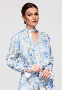 Mujer que lleva una blusa floral azul y blanca con lazo en el cuello y botones de perla, ajustando la cinta de la manga sobre un fondo liso.