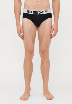 Personne mince debout pieds nus portant un slip noir avec une taille blanche portant l'inscription "SEX®" sur un fond blanc uni.