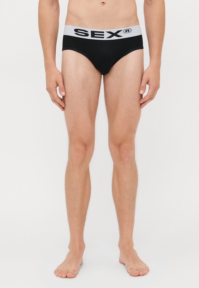 Schlanke Person, die barfuß steht und schwarze Shorts mit einem weißen Bund trägt, auf dem "SEX®" steht, vor einem schlichten weißen Hintergrund.