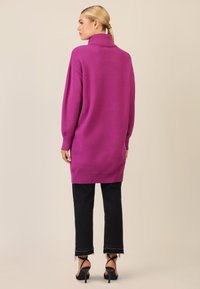 Fuchsiefarbenes Oversized-Pullover mit hohem Kragen und Rippensaum, getragen über schwarzen, cropped Jeans mit ausgefranstem Saum und schwarzen Riemchen-Heels.
