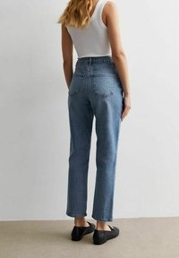 Jeans de mezclilla azul de cintura alta con pierna recta, que cuentan con bolsillos traseros y un lavado claro. Combinados con una camiseta sin mangas blanca ajustada y zapatos negros.