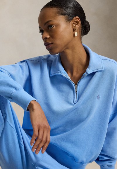 Ανοιχτό μπλε φούτερ με φερμουάρ και γιακά· απαλό ύφασμα· λογότυπο στο στήθος· άνετη γραμμή με μακριά μανίκια.