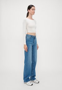 Weißes Langarm-Crop-Top, hochgeschnittene blaue Jeans und weiße Sneakers. Die Jeans sind locker sitzend mit einem geraden Bein-Design.