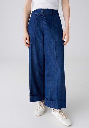 Weit geschnittene dunkelblaue Jeanshose mit großen vorderen Taschen, umgeschlagenen Säumen und gebundenem Taillengürtel, kombiniert mit weißen Turnschuhen und einem weißen ärmellosen Oberteil.