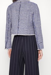 Kort jacka i blått och vitt strukturerat tyg med fransade kanter. Har långa ärmar och en öppen ryggdesign. Matchas med randiga byxor.