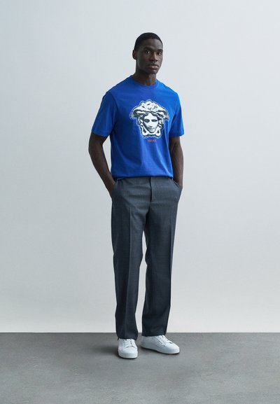 Modré bavlnené tričko s grafickou potlačou Medusy, skombinované so sivými spoločenskými nohavicami a bielymi teniskami, vytvárajúce moderný ležérny vzhľad.