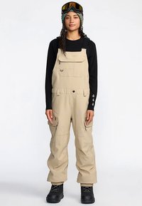 Beige snow overalls made of durable material, featuring multiple pockets, adjustable straps, and a relaxed fit, paired with black footwear.