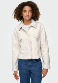 Donna con capelli ricci che indossa un giubbotto sherpa bianco e jeans blu, in piedi con le mani rilassate su uno sfondo neutro.