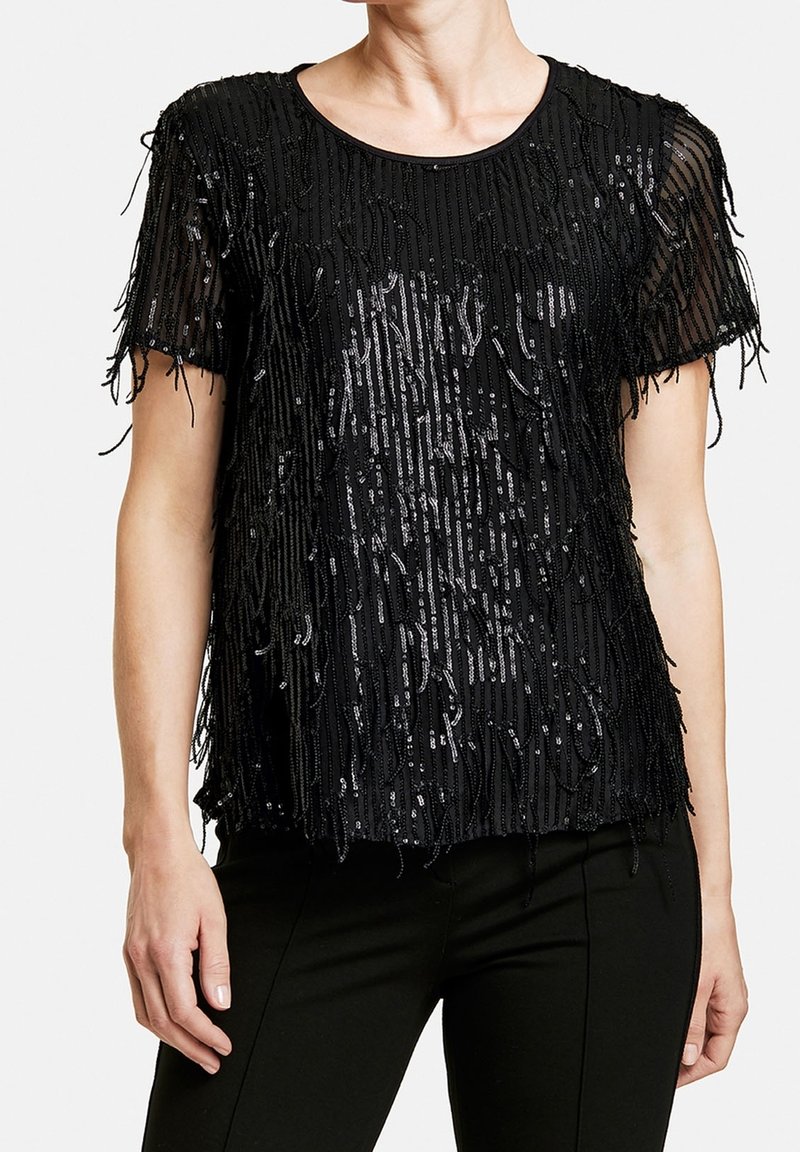 Top nero con maniche corte, caratterizzato da dettagli in frange e paillettes. Il tessuto trasparente aggiunge texture al design. Orlo piatto senza vita definita.