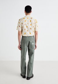 Short-sleeve shirt with fruit pattern in beige, paired with olive green pants featuring rear pockets and contrasting stitching. Black boots visible.
