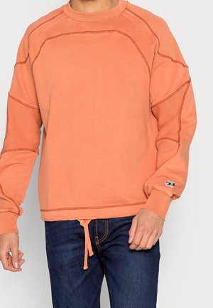 Osoba ubrana w koralową bluzę z długim rękawem z widocznymi przeszyciami, w połączeniu z ciemnoniebieskimi jeansami.