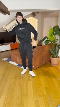 Moletom preto e calças de moletom com detalhes de logotipo; segurando uma caixa castanha da Zalando. O quarto apresenta uma planta verde e um sofá alaranjado texturizado.