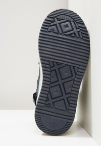 Suela de zapato de goma negra con un patrón de agarre texturizado que presenta formas geométricas y un emblema de la marca. Construcción sólida con material duradero.