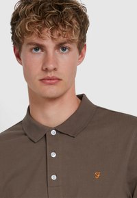 Braunes Poloshirt mit Kragen, versehen mit einem orangefarbenen Logos auf der Brust und weißen Knöpfen. Der Stoff wirkt strukturiert und tailliert.