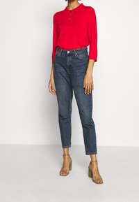 Rotes langärmliges Poloshirt mit Kragen und Knopfleiste, kombiniert mit hochgeschnittenen dunkelblauen Jeans und grünen Blockabsatzsandalen.