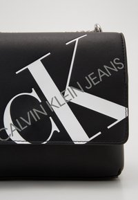 Černá kožená taška s velkým bílým logem "CK" a šedým textem "Calvin Klein Jeans" napříč přední klopou, zdobená stříbrným řetězem.