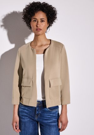 Veste légère beige avec un col rond, deux poches avant, des manches trois-quarts et une texture lisse, associée à un débardeur blanc et un jean bleu.