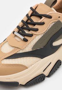 Baskets épaisses avec mesh beige, panneaux en cuir marron clair et noir, lacets noirs avec embout argenté, et semelle épaisse crème avec crampons noirs.