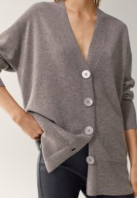 Szary, oversize kardigan wykonany z miękkiego materiału dzianinowego. Posiada głęboki dekolt w kształcie V, cztery duże guziki oraz ściągacze przy mankietach i dolnej krawędzi.