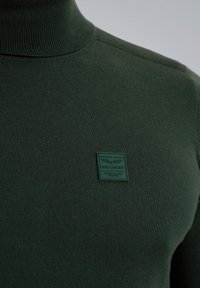 Dunkelgrüner Rollkragenpullover aus Strickstoff, mit einer strukturierten Oberfläche und einem quadratischen grünen Etikett mit geprägtem Text.