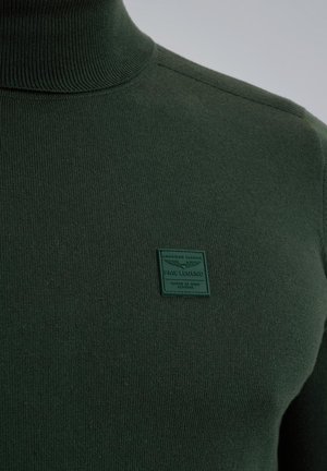 Mørkegrøn turtleneck sweater lavet af strikket stof, med en struktureret overflade og et firkantet grønt mærke med prægede bogstaver.