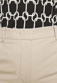 Beige byxor med en mjuk textur, med rak passform, bälteshällor och minimala sömnadsdetaljer. Bakgrunden visar en mönstrad topp.