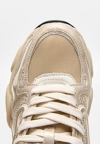 Béžová teniska s horní částí z síťoviny, semišovými překryvy a bílými tkaničkami uvázanými ve standardním zkříženém vzoru.