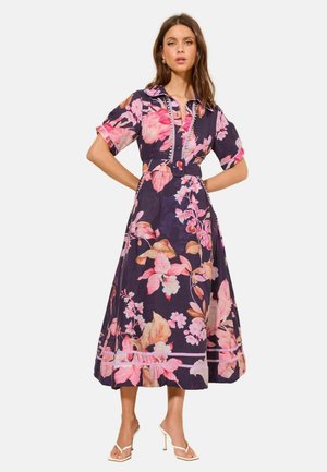 Žena v tmavofialových midi šatách s veľkým ružovým kvetinovým vzorom, so zapínaním v páse, krátkymi rukávmi a bielymi sandálmi na vysokom podpätku.