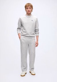 Mand står iført lysegrå sweatshirt og matchende joggingbukser med hvide sneakers mod en ensfarvet hvid baggrund.
