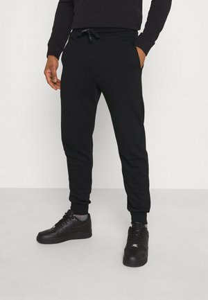 Person iført sorte joggingbukser, sorte sneakers og en sort sweatshirt står med den ene hånd i lommen mod en ensfarvet baggrund.