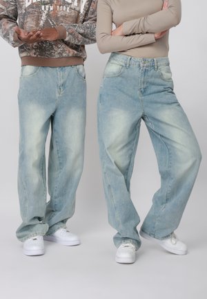 Deux personnes portant des jeans larges bleu clair et des baskets blanches, l'une avec un sweat à capuche camouflage et l'autre un haut à manches longues beige, les bras croisés.