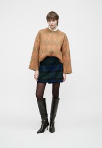 Pull surdimensionné orangé à motifs, jupe écossaise bleu-vert, collants noirs et bottes noires brillantes jusqu'aux genoux. Textures lisses, design moderne.