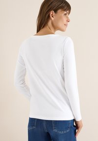 Weißes Langarmhemd mit rundem Ausschnitt, glatter Textur, figurbetontem Schnitt, kombiniert mit blauen Jeans, minimalistisches Design.