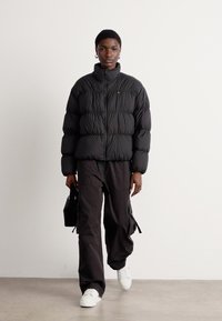 Svart pufferjakke med quiltet design, glidelåslukking og høy krage. Kombinert med løstsittende svarte cargobukser og hvite joggesko.